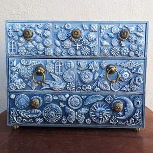Puede incluir: Una caja de madera azul para joyas con cuatro cajones. Cada caj&oacute;n est&aacute; decorado con un dise&ntilde;o diferente de flores, mariposas y otros motivos inspirados en la naturaleza. La caja tiene tiradores de lat&oacute;n y un acabado desgastado.