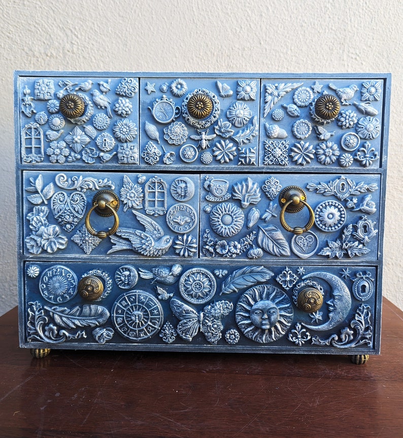 Puede incluir: Una caja de madera azul para joyas con cuatro cajones. Cada caj&oacute;n est&aacute; decorado con un dise&ntilde;o diferente de flores, mariposas y otros motivos inspirados en la naturaleza. La caja tiene tiradores de lat&oacute;n y un acabado desgastado.
