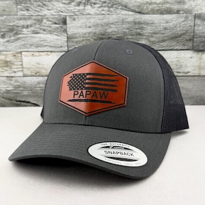 May include: A charcoal gray snapback hat with a black mesh back and a brown hexagonal leather patch on the front. The patch features a distressed American flag graphic and the text PAPAW. A circular sticker on the brim reads THE AUTHENTIC YUPOONG SNAPBACK.