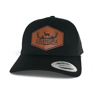 May include: A black snapback cap featuring a brown hexagonal patch on the front. The patch displays a black graphic of a deer, antlers, trees, and the word GRANDPA above two crossed rifles. A circular sticker on the brim reads THE AUTHENTIC SNAPBACK.