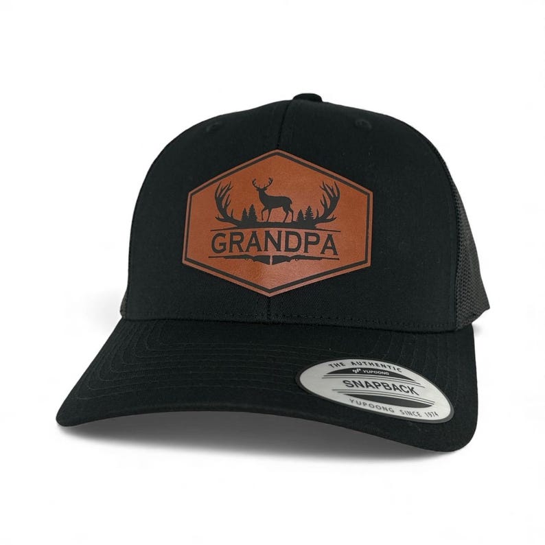 May include: A black snapback cap featuring a brown hexagonal patch on the front. The patch displays a black graphic of a deer, antlers, trees, and the word GRANDPA above two crossed rifles. A circular sticker on the brim reads THE AUTHENTIC SNAPBACK.