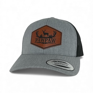May include: A heather gray baseball cap with a black mesh back and a brown hexagonal patch on the front. The patch features a black silhouette of a deer with antlers above the text PAWPAW. A round sticker on the brim reads The Authentic Yupoong Snapback.