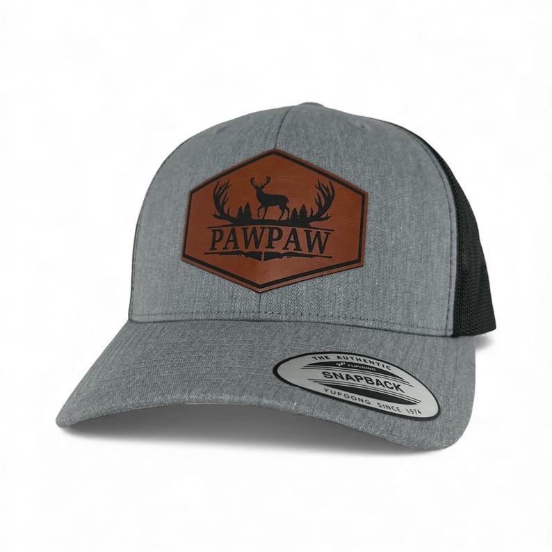 May include: A heather gray baseball cap with a black mesh back and a brown hexagonal patch on the front. The patch features a black silhouette of a deer with antlers above the text PAWPAW. A round sticker on the brim reads The Authentic Yupoong Snapback.