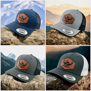 May include: Four snapback caps with mesh backs and leather-like hexagonal patches. The patches feature a deer silhouette, antlers, and the text PAPAW, DAD, GRANDPA, or POPS. The caps are navy, heather gray, dark gray, and charcoal gray.