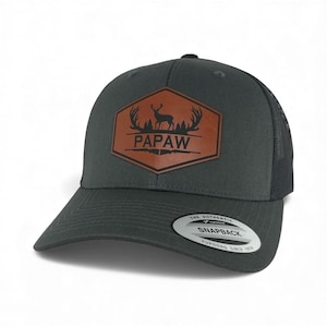 May include: A charcoal gray snapback cap featuring a brown hexagonal leather patch on the front. The patch displays a black silhouette of a deer with antlers, pine trees, and the text PAPAW. A round sticker on the brim reads THE AUTHENTIC SNAPBACK.