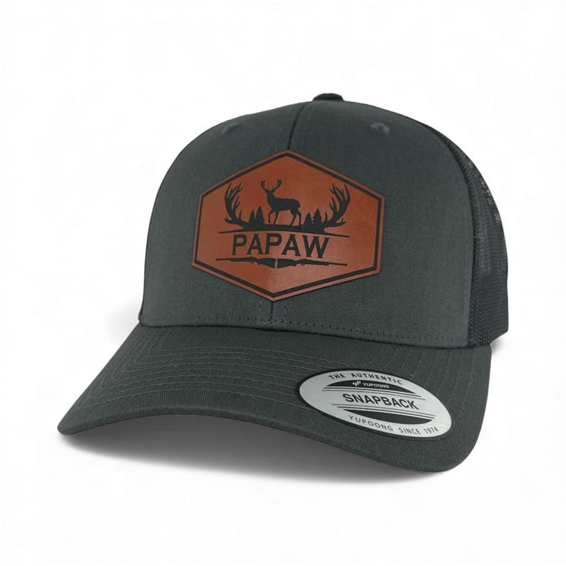 May include: A charcoal gray snapback cap featuring a brown hexagonal leather patch on the front. The patch displays a black silhouette of a deer with antlers, pine trees, and the text PAPAW. A round sticker on the brim reads THE AUTHENTIC SNAPBACK.