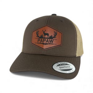 May include: A brown snapback cap with a tan mesh back. The front features a hexagonal leather patch with a black graphic of a deer, antlers, and trees above the text PAPAW, with two rifles below. A circular sticker is on the flat brim.