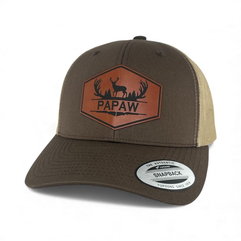 May include: A brown snapback cap with a tan mesh back. The front features a hexagonal leather patch with a black graphic of a deer, antlers, and trees above the text PAPAW, with two rifles below. A circular sticker is on the flat brim.