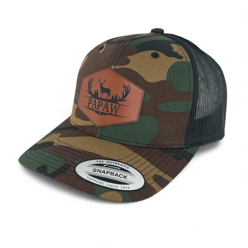 May include: A camouflage print snapback hat with a black mesh back. A brown hexagonal patch on the front features a black silhouette of a deer with antlers and the text PAPAW. A circular sticker on the brim reads THE AUTHENTIC YUPOONG SNAPBACK.