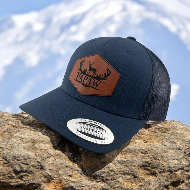 May include: A navy blue snapback cap featuring a brown leather-like hexagonal patch on the front. The patch displays a black silhouette of a deer with antlers and the text PAPAW. The cap has a mesh back and a sticker on the brim reading THE AUTHENTIC YUPOONG SNAPBACK YUPOONG SINCE 1974.