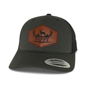 May include: A charcoal gray snapback cap featuring a brown hexagonal leather patch on the front. The patch displays a black silhouette of a deer with antlers above the text PAPPY. The cap has a black mesh back and a sticker on the brim.