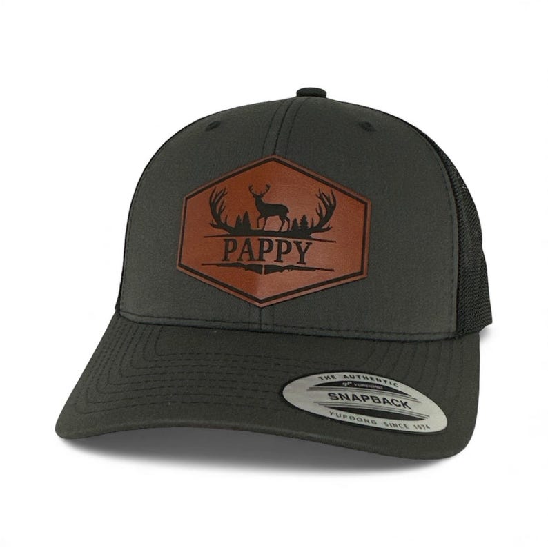 May include: A charcoal gray snapback cap featuring a brown hexagonal leather patch on the front. The patch displays a black silhouette of a deer with antlers above the text PAPPY. The cap has a black mesh back and a sticker on the brim.