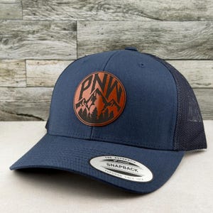 May include: A navy blue snapback trucker hat featuring a circular brown leather patch on the front. The patch displays a black mountain range silhouette, pine trees, and the letters PNW. A sticker on the brim reads The Authentic Yupoong Snapback.