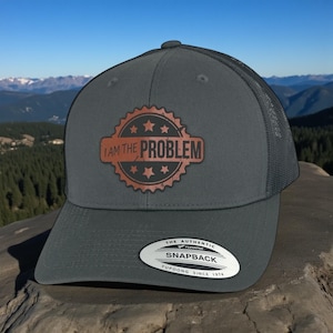 May include: A charcoal grey trucker hat with a mesh back and a snapback closure. The front features a circular brown patch with a serrated edge, four stars, and the text "I AM THE PROBLEM" in black. An oval sticker on the brim reads "THE AUTHENTIC YUPOONG SNAPBACK YUPOONG SINCE 1974".