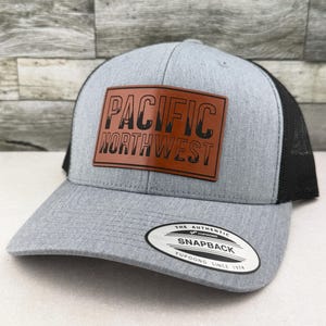May include: A heather gray baseball cap with a black mesh back and a brown rectangular leather patch on the front. The patch features the black text PACIFIC NORTHWEST. A circular black and white sticker on the brim reads THE AUTHENTIC YUPOONG SNAPBACK.