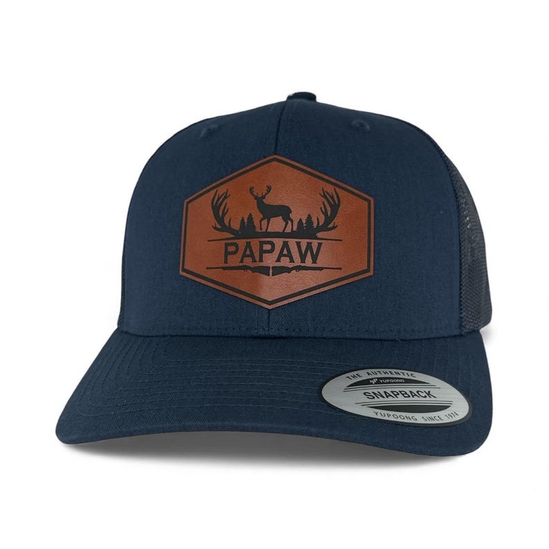 May include: A navy blue snapback cap featuring a brown hexagonal leather patch on the front. The patch displays a black graphic of a deer, antlers, and the text PAPAW. A circular sticker on the brim reads The Authentic Yupoong Snapback.