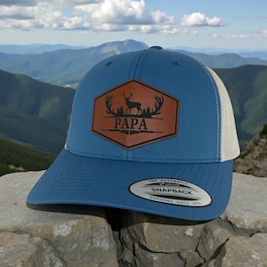 May include: A blue snapback cap with a beige mesh back and a brown hexagonal leather patch on the front. The patch features a black graphic of a deer silhouette, antlers, and pine trees with the text PAPA. A sticker is on the flat brim.