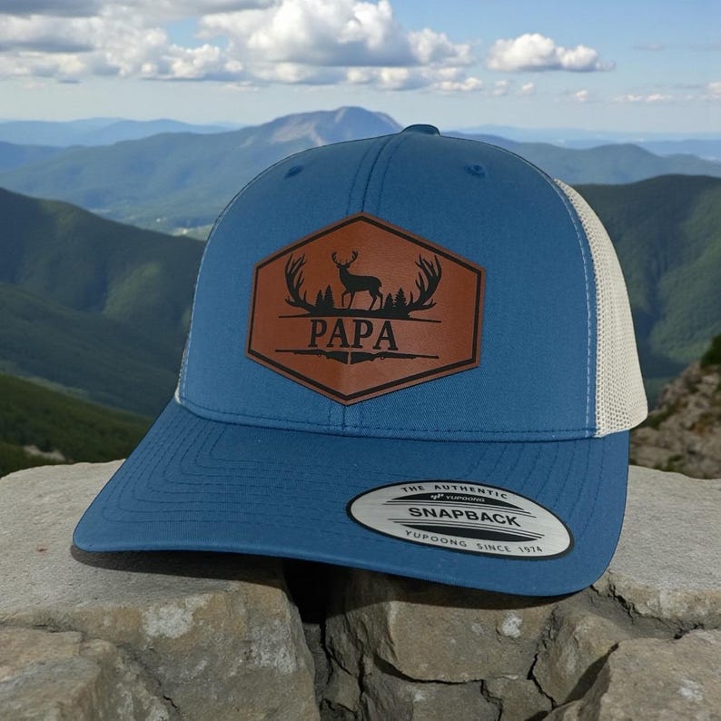 May include: A blue snapback cap with a beige mesh back and a brown hexagonal leather patch on the front. The patch features a black graphic of a deer silhouette, antlers, and pine trees with the text PAPA. A sticker is on the flat brim.