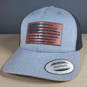 May include: A heather grey baseball cap with a black mesh back and a brown rectangular leather patch featuring a distressed American flag design. The brim displays a circular sticker with the text The Authentic Yupoong Snapback Since 1974.