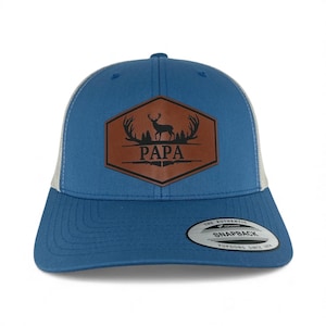 May include: A blue snapback cap with a white mesh back and a brown hexagonal leather patch on the front. The patch features a black graphic of a deer, antlers, and trees above the text PAPA, with a rifle silhouette below the word.