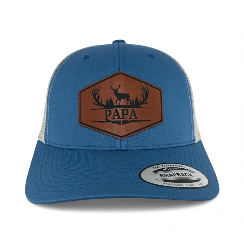 May include: A blue snapback cap with a white mesh back and a brown hexagonal leather patch on the front. The patch features a black graphic of a deer, antlers, and trees above the text PAPA, with a rifle silhouette below the word.