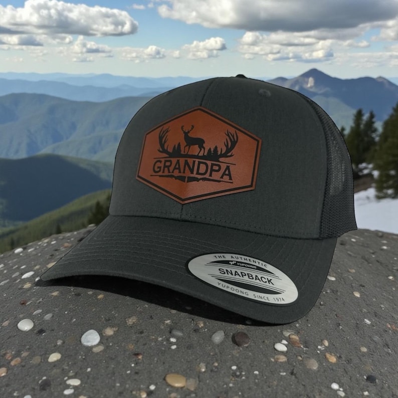 May include: A charcoal gray trucker hat with a mesh back and a brown leather-like hexagonal patch on the front. The patch features a black silhouette of a deer with antlers and the word GRANDPA printed in bold black capital letters.