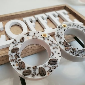 Könnte beinhalten: Zwei runde Ohrringe aus weißem Harz mit einem Kaffeekorn-Design. Die Ohrringe haben einen goldenen Akzent.