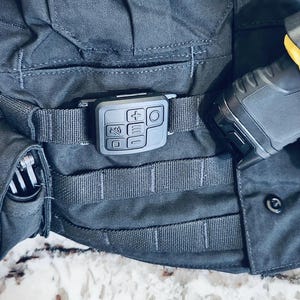 May include: A black tactical vest with a black button and a black device with a small screen and buttons attached to the front. The device has a circular button on the top right corner.