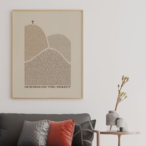 May include: A beige framed print with a black cross at the top and the text "SERMON ON THE MOUNT" at the bottom. The print features a stylized image of two mountains made up of text.