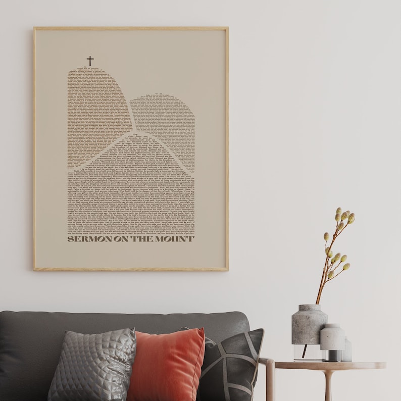 May include: A beige framed print with a black cross at the top and the text "SERMON ON THE MOUNT" at the bottom. The print features a stylized image of two mountains made up of text.