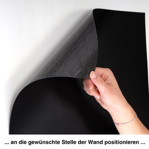 May include: A black magnetic sheet being applied to a white wall. The sheet is being held in place by a hand.
