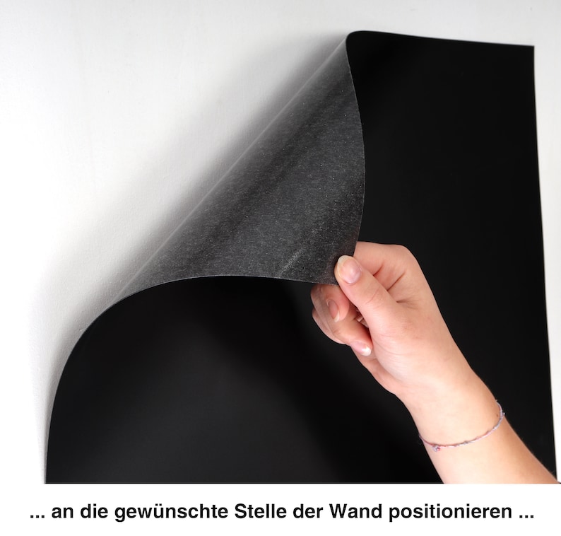 May include: A black magnetic sheet being applied to a white wall. The sheet is being held in place by a hand.