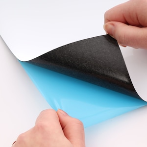 Puede incluir: Una pizarra blanca magn&eacute;tica de borrado en seco con un respaldo negro y una capa adhesiva azul. La capa adhesiva se est&aacute; despegando para revelar el adhesivo.