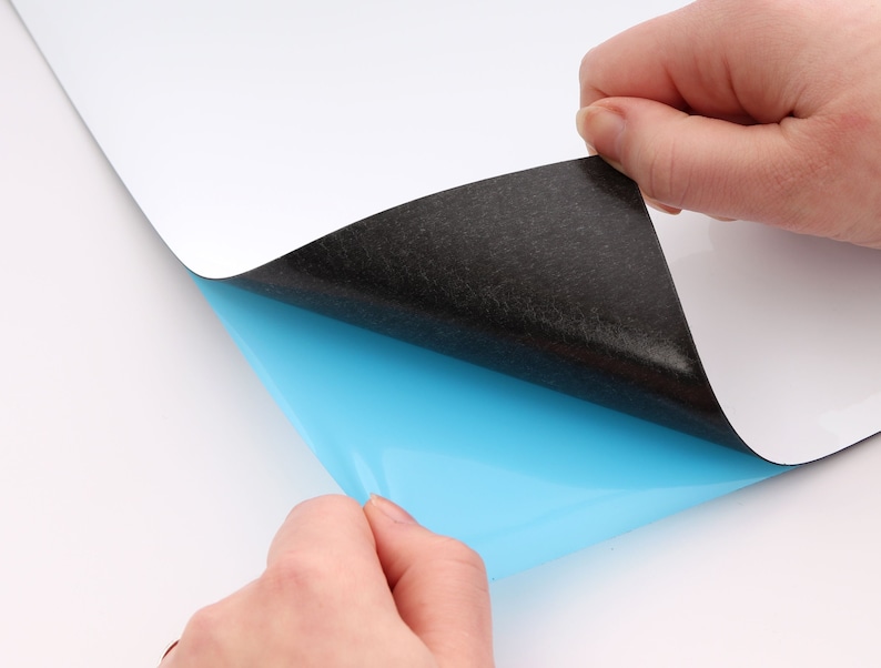 Puede incluir: Una pizarra blanca magn&eacute;tica de borrado en seco con un respaldo negro y una capa adhesiva azul. La capa adhesiva se est&aacute; despegando para revelar el adhesivo.
