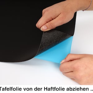 May include: A hand peeling back a black chalkboard sticker from a blue adhesive backing. The text "Tafelfolie von der Haftfolie abziehen..." is visible at the bottom of the image.