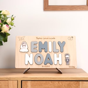 May include: A wooden puzzle with the names "EMILY" and "NOAH" in gray and white letters. The puzzle includes illustrations of a bride and groom, doves, and wedding rings. The date "28.03.2025" is also displayed.