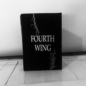 Könnte beinhalten: Ein schwarzes Buch mit dem Titel "FOURTH WING" in weißen Lettern. Der Buchumschlag zeigt einen stilisierten Blitz und ein astähnliches Design.
