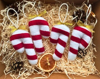 Puede incluir: Cuatro adornos de bastón de caramelo con rayas rojas y blancas, cada uno con un lazo de cinta dorada y una cuerda blanca para colgar. Los adornos están colocados en paja con piñas y una rodaja de naranja seca.