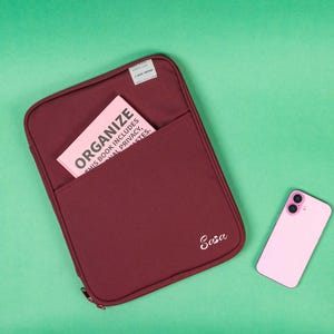 May include: A burgundy organiser case with a pocket holding a pink book that reads "ORGANIZE". A pink smartphone is next to the case. The case has the name "Sasa" embroidered on it.