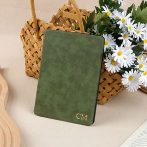 Könnte beinhalten: Ein grünes Lederjournal mit goldenen, geprägten Initialen "CM" auf dem vorderen Umschlag. Das Journal liegt auf einer hellbraunen Oberfläche neben einem geflochtenen Korb, der mit weißen Gänseblümchen gefüllt ist.