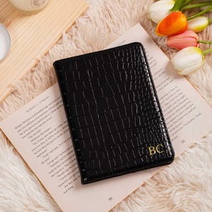 May include: Black faux-leather wallet with a crocodile texture, featuring gold-colored initials "BC". The wallet is resting on a book with text and is surrounded by a soft, fluffy surface and flowers.