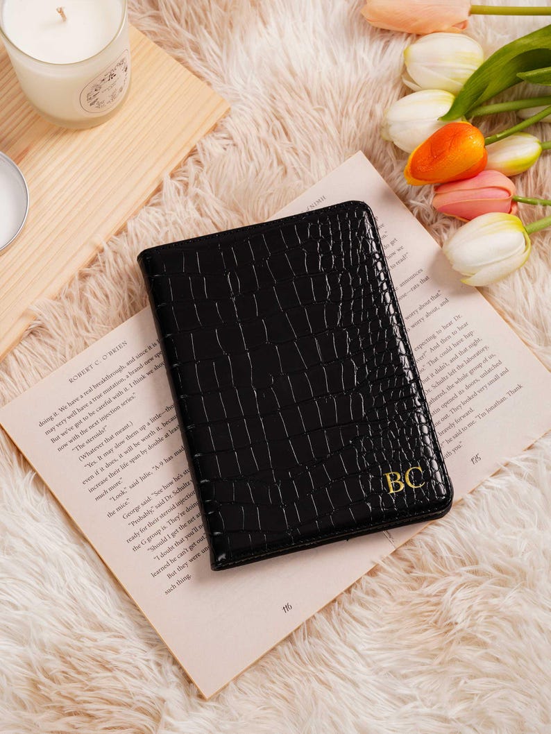 May include: Black faux-leather wallet with a crocodile texture, featuring gold-colored initials "BC". The wallet is resting on a book with text and is surrounded by a soft, fluffy surface and flowers.