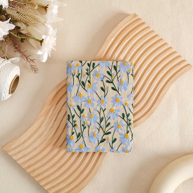 Pode incluir: Uma pequena capa de caderno bordada com flores azul claro e verde. A capa &eacute; feita de um tecido macio e natural e apresenta um padr&atilde;o floral delicado.