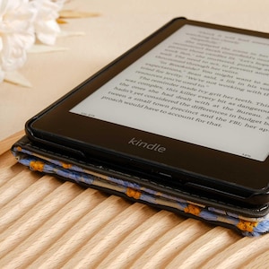 Pode incluir: Um leitor de e-books Kindle preto com uma capa estampada com flores azuis e amarelas. A tela exibe texto de um livro.