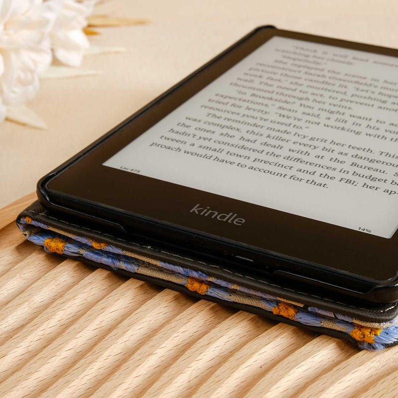 Pode incluir: Um leitor de e-books Kindle preto com uma capa estampada com flores azuis e amarelas. A tela exibe texto de um livro.