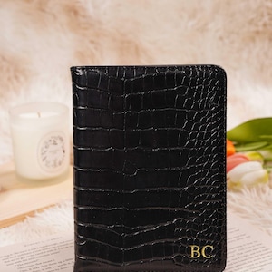 May include: Black faux-leather passport holder with a crocodile skin texture. The holder features gold-colored initials "BC" on the lower right corner. The background includes a candle, flowers, and a book.