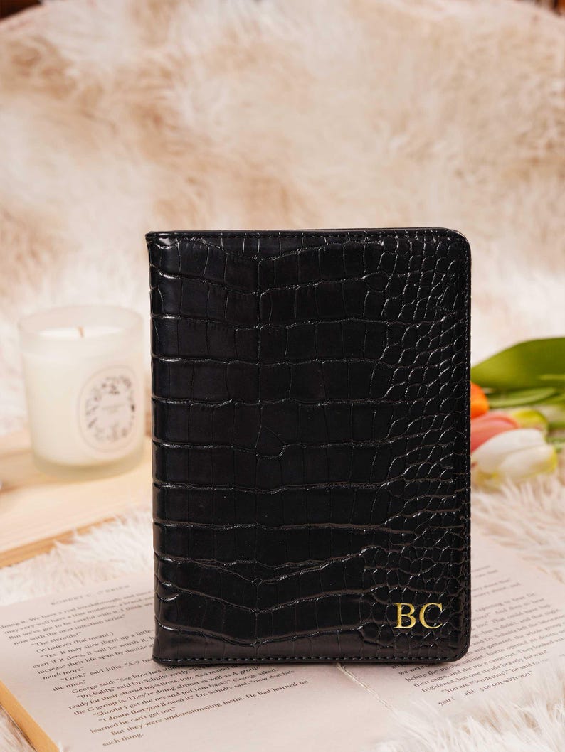May include: Black faux-leather passport holder with a crocodile skin texture. The holder features gold-colored initials "BC" on the lower right corner. The background includes a candle, flowers, and a book.