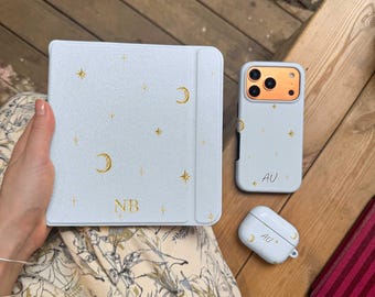 May include: A light blue e-reader cover with gold moon and star accents and the initials NB, a matching light blue phone case with the initials AU, and a matching light blue wireless earbud case with the initials AU, all featuring gold celestial motifs.