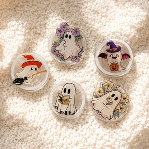 May include: Five circular phone grips featuring cartoon ghost illustrations. One ghost wears a witch hat on a broom, one is surrounded by purple flowers, one has bat wings holding a pumpkin, one reads a book titled Fourth Wing, and one holds yellow flowers.
