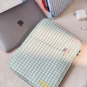 May include: A sage green and white gingham laptop sleeve with a zippered closure, a front pocket, and the name Sasa embroidered in yellow. A small rectangular tag on the upper right corner features the text A GOOD PARTNER in black lettering.